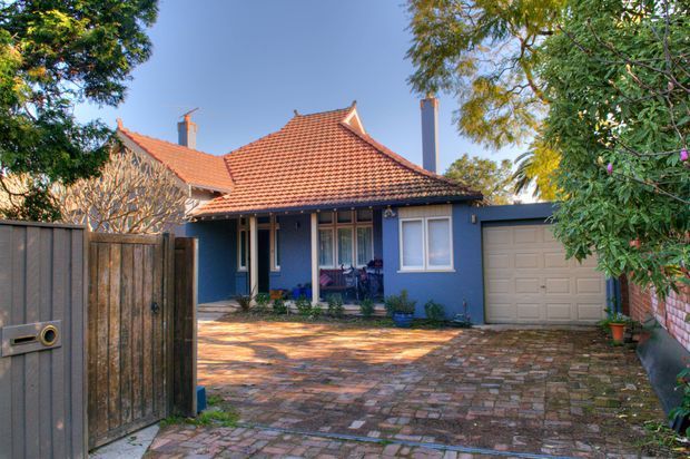 Street-facing view of a traditional single-storey brick-paved residence with a terracotta tiled hipped roof, blue-painted facade, white-framed windows, covered entry porch, and an attached garage. Mature trees and garden planting frame the driveway and front courtyard.