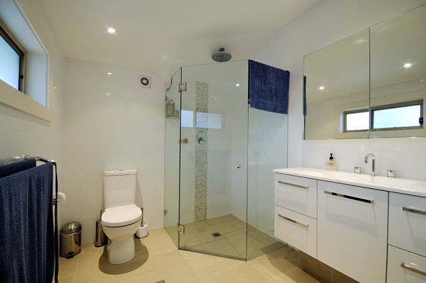 Bright contemporary bathroom with a frameless glass walk-in shower, wall-mounted white vanity, large mirrored cabinet, toilet, towel rail, and high horizontal window. The room uses a clean white palette with beige floor tiles and a blue tiled shower feature.