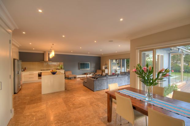 Open-plan residential interior combining a contemporary kitchen, dining area, and living room. The space has warm polished stone-look flooring, white cabinetry with a central island, a dark grey feature wall, recessed ceiling lighting, a grey sectional sofa, and large windows opening toward a bright garden or courtyard. A timber dining table with upholstered chairs occupies the foreground.