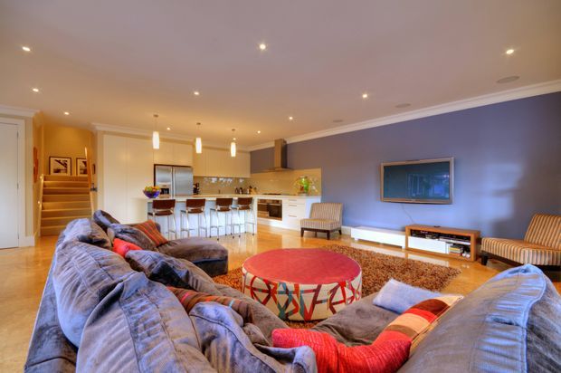 Open-plan living room and kitchen with a large sectional sofa in the foreground, colourful round coffee table, wall-mounted television, timber flooring, and a compact white kitchen with island seating. The room has recessed ceiling lighting, pendant lights over the island, a blue feature wall, and warm neutral finishes.