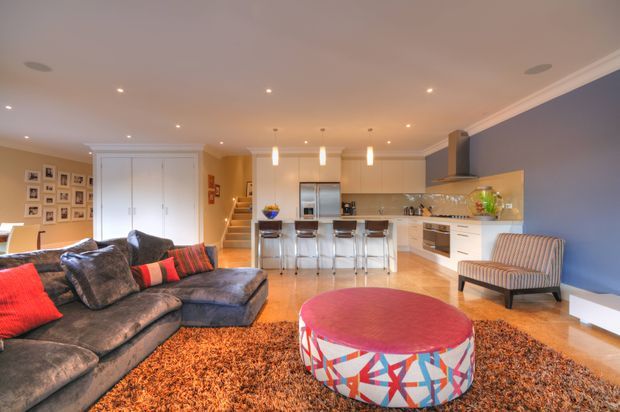 Open-plan contemporary living area with a large dark sectional sofa, patterned red and black cushions, a vibrant round upholstered coffee table, and a textured orange-brown rug. The adjoining kitchen has white cabinetry, a long island with bar seating, stainless-steel appliances, pendant lights, and a warm timber floor. A deep blue feature wall, cream walls, recessed lighting, framed family photographs, and a staircase complete the inviting space.