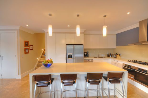 Bright, spacious contemporary kitchen viewed straight on, featuring a large white island with four bar stools, extensive white built-in cabinetry, stainless-steel French-door refrigerator, integrated appliances, a gas cooktop and rangehood. Warm beige walls and flooring are balanced by a dark blue-grey feature wall, with three pendant lights above the island and a staircase visible beyond.
