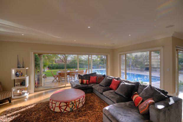 Spacious, warmly lit living room with a large dark grey sectional sofa, colourful patterned cushions, a round upholstered coffee table, and a deep shag rug. Full-height sliding glass doors and windows open the room visually to a timber dining deck and swimming pool beyond. Neutral beige walls, recessed ceiling lighting, timber flooring, and a built-in media area create a relaxed contemporary family entertaining space.