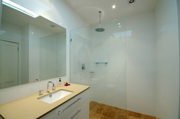Bright contemporary bathroom featuring a large frameless glass walk-in shower, ceiling-mounted rain showerhead, wall-mounted shower controls, floating glass shelf, and a long pale stone vanity with integrated white basin and chrome tapware. The room has glossy white wall and ceiling finishes, recessed lighting, mirrored surfaces, and warm brown tiled flooring.