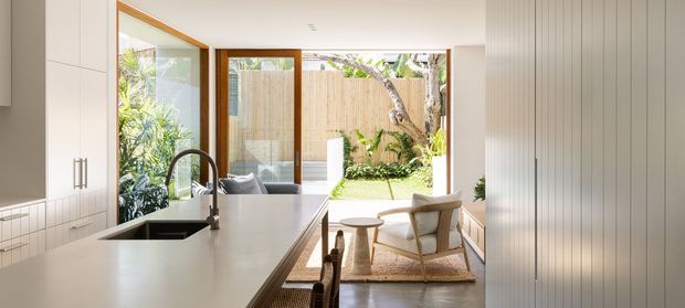 Bright open-plan kitchen with a long light benchtop and island sink, white cabinetry, timber-framed glazing and a glazed timber door opening to a landscaped courtyard. A small seating area with pale upholstered chairs is visible beyond the kitchen, with abundant natural light and a warm, restrained material palette.