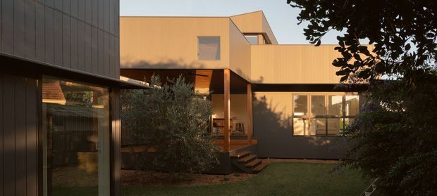 Exterior view of a contemporary multi-volume residence arranged around a landscaped courtyard. The building has warm timber-toned cladding, dark vertical-panel cladding on the foreground volume, large glazed openings, projecting upper-level forms, and a sheltered entry reached by steps. Mature planting and lawn frame the scene in warm late-day light.