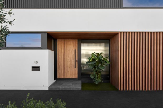 Contemporary residential front entry with a crisp white rendered wall, dark charcoal roof and paving, warm timber-clad recessed doorway, vertical timber screening, and a narrow planted entry court. The facade combines clean horizontal lines with a strongly framed timber portal and integrated mailbox.