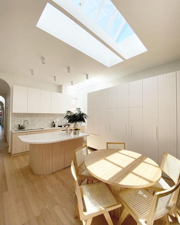 Bright open-plan kitchen and dining area with a large skylight, white full-height cabinetry, pale timber flooring, a curved island with a vertical timber-slatted base and light stone benchtop, and a round timber dining table surrounded by woven cane chairs. The space has abundant natural light and a clean, warm minimalist aesthetic.