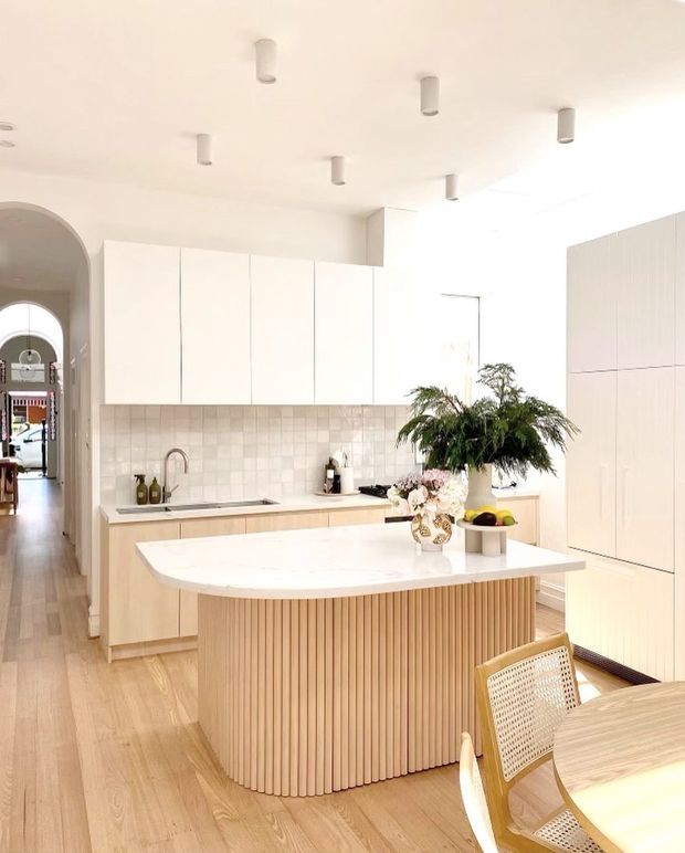 Bright, contemporary open-plan kitchen with a large curved island featuring a white stone benchtop and vertical timber slat detailing. The room has pale timber flooring, light timber lower cabinetry, white handleless upper cabinetry, a softly patterned tiled splashback, integrated tall storage, recessed and surface-mounted ceiling lighting, and an arched hallway opening. A dining table and cane-backed chairs are visible in the foreground.