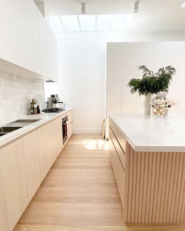 Bright contemporary kitchen with a pale timber palette, white cabinetry, integrated appliances, tiled splashback, and a large central island. The island features a white stone benchtop and a vertically fluted timber-clad base. A skylight or translucent roof panel brings abundant natural light into the space, complemented by simple cylindrical ceiling lights and restrained floral styling.