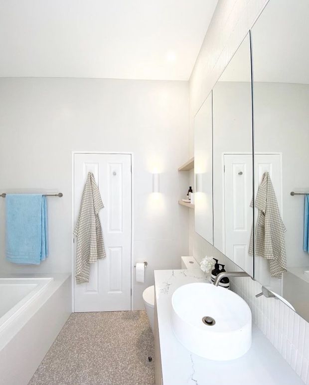 Bright, compact contemporary bathroom with a white freestanding vessel basin on a white marble-look vanity, large mirrored wall cabinetry, bathtub, toilet, towel rails, and soft wall lighting. The room uses a clean white palette with subtle grey veining and a textured neutral floor.