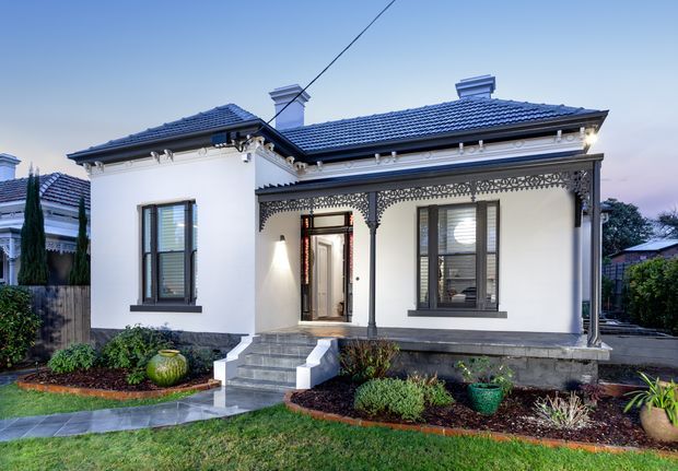 Front exterior view of a compact heritage-style single-storey house with white rendered walls, a dark tiled hipped roof, decorative lacework verandah detailing, timber-framed windows, and a raised entry porch with steps. The landscaped front garden and path are visible, with neighbouring houses in the background.