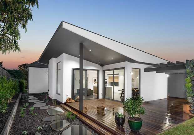 Contemporary single-storey house exterior at dusk, featuring crisp white rendered walls, extensive floor-to-ceiling glazing, a dramatic asymmetrical pitched roof, covered timber decking and an adjoining pergola-like outdoor area. A landscaped stepping-stone path leads toward the entrance, with warm interior lighting visible through the windows.