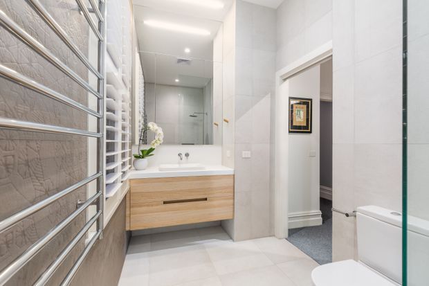 Bright contemporary bathroom with large light-grey tiled walls and floor, a floating light timber vanity with integrated white basin, mirrored cabinetry, chrome heated towel rail, frameless glass shower screen and white toilet. A doorway opens to an adjacent carpeted room.