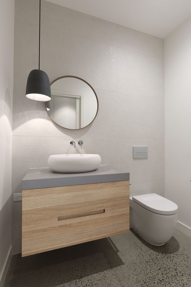 Compact contemporary bathroom with a wall-mounted light-wood vanity, grey stone-look benchtop, white vessel basin, round framed mirror, wall-hung toilet and textured pale wall tiles. A dark pendant light provides a refined contrast, while the speckled grey floor adds visual texture.