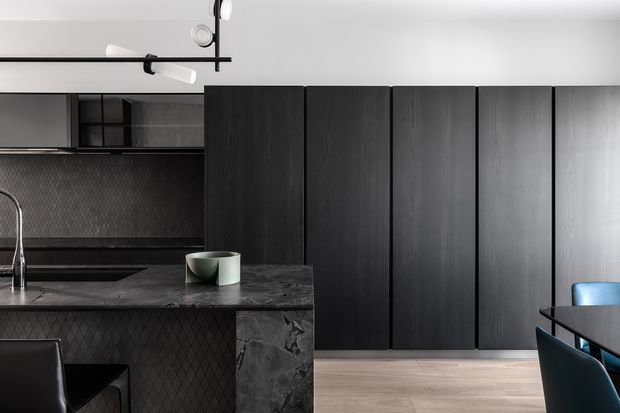 A dark, contemporary kitchen with tall charcoal cabinetry, a black stone island and benchtop, textured dark backsplash, integrated cooktop and sink, and a minimalist black track light. Pale timber flooring and vivid blue dining chairs provide contrast.