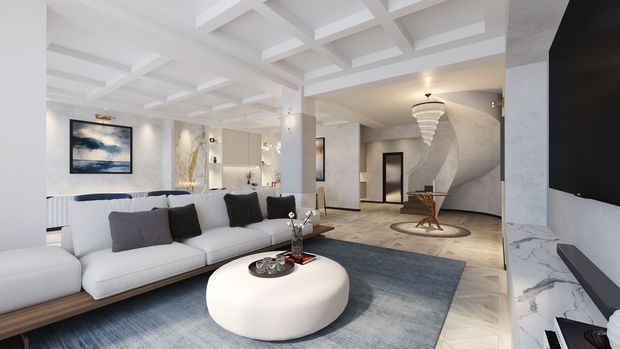 A spacious, refined open-plan living room with a large white sectional sofa, round upholstered coffee table, blue-grey area rug, marble-clad fireplace or media wall, coffered ceiling, sculptural pendant light, and pale wood flooring. The room includes a small dining or reading area in the background and contemporary artwork and decor.