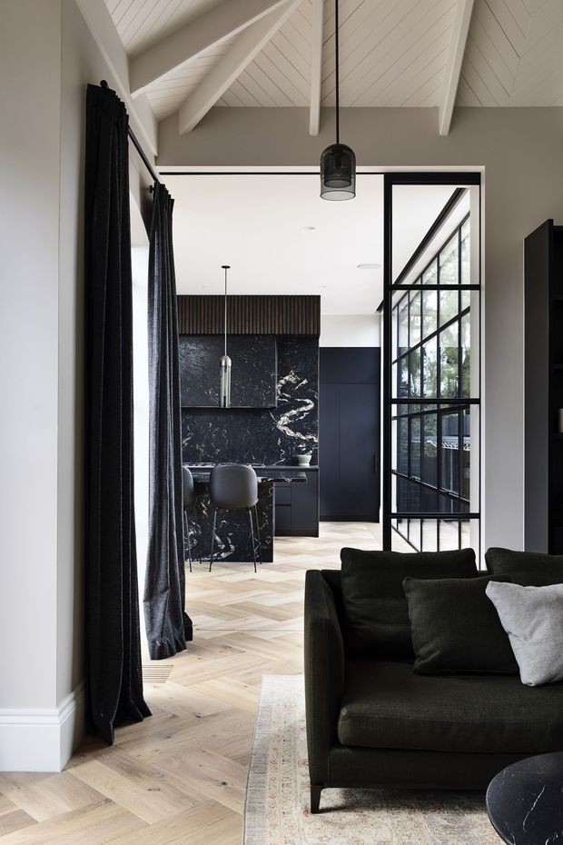 A refined open-plan interior viewed from a living room toward a dark, marble-featured kitchen. The room has pale herringbone timber flooring, charcoal curtains, a deep green-black sofa, black-framed internal glazing, and a dramatic vaulted ceiling with white painted exposed beams and panelling. A black pendant light hangs centrally, while the kitchen includes dark cabinetry, a stone island, fluted timber detailing, and bar stools.