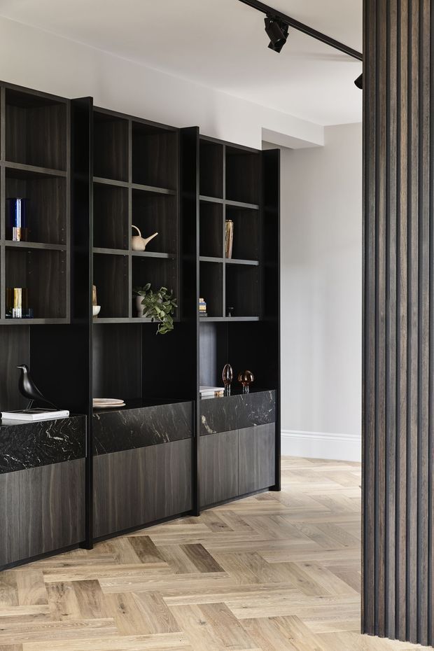 A refined interior featuring a full-height dark built-in shelving and storage wall with open display niches, dark timber cabinetry, and veined black stone benchtop sections. The shelving is styled with minimal books, ceramics, and decorative objects. A vertical dark slatted screen frames the right side, while pale herringbone timber flooring and white walls create contrast. Black track lighting runs across the ceiling.