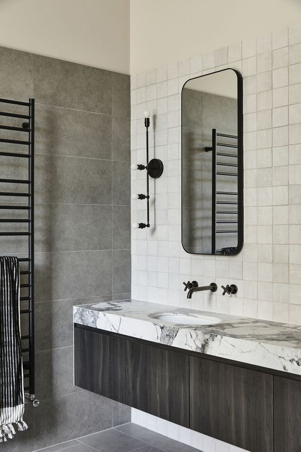 Contemporary bathroom featuring a wall-mounted dark timber vanity with an integrated white-and-grey veined stone benchtop and inset basin. The walls combine large grey stone-look tiles with small white square tiles, complemented by matte black tapware, a rounded-rectangle black-framed mirror, and a black heated towel rail.