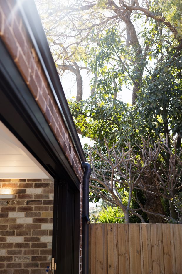 View of a residential exterior corner featuring exposed red-brown brick walls, dark metal roof and guttering, and a partially visible covered opening. A timber paling fence and dense mature garden vegetation frame the property, with bright daylight filtering through the trees.