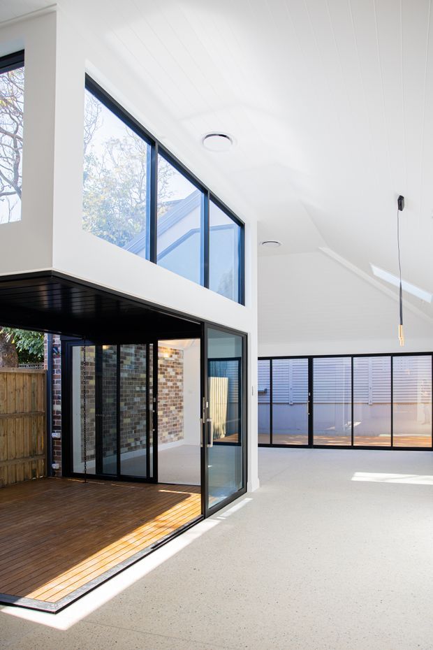 Bright, double-height contemporary interior with extensive black-framed glazing opening onto a timber deck. The space features a white-lined ceiling, polished light concrete floor, exposed brickwork visible through the glazed opening, and strong indoor-outdoor connection. A pendant light is suspended from the ceiling, with upper-level windows and a broad bank of louvred glazing adding to the airy architectural character.
