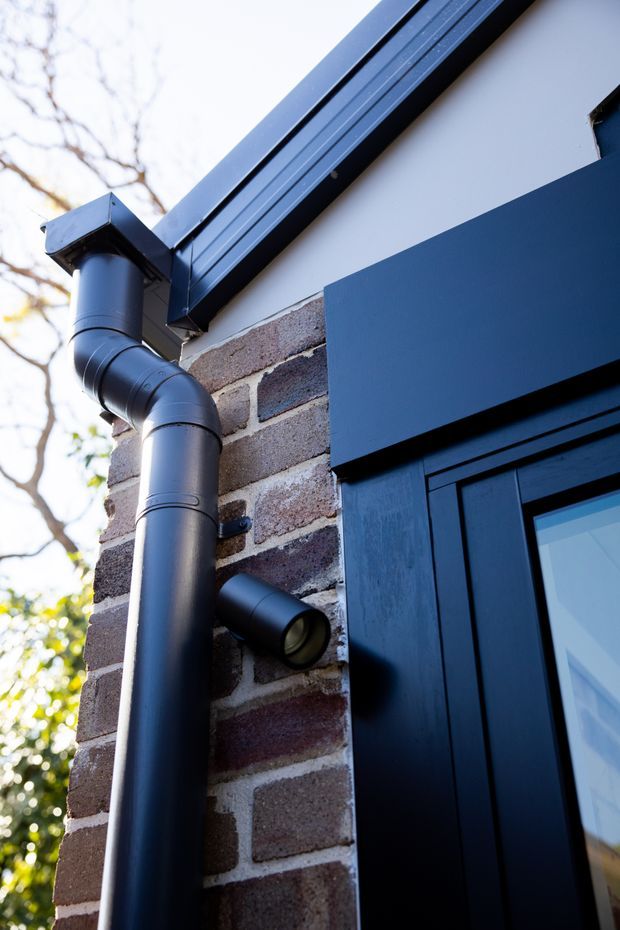 Close-up exterior detail of a dark-painted brick facade, featuring a black downpipe and gutter, dark blue-black door and trim, and a small cylindrical wall light beside the entry.