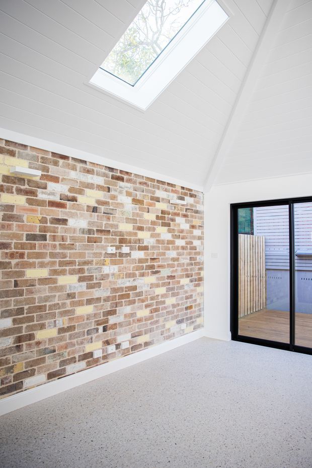 Bright, largely empty interior room with a high white-lined pitched ceiling and a prominent skylight. One wall features exposed multicoloured brickwork, while black-framed sliding glass doors open to a small timber deck or courtyard. The floor is a light speckled hard surface, and the space has a clean, contemporary feel.
