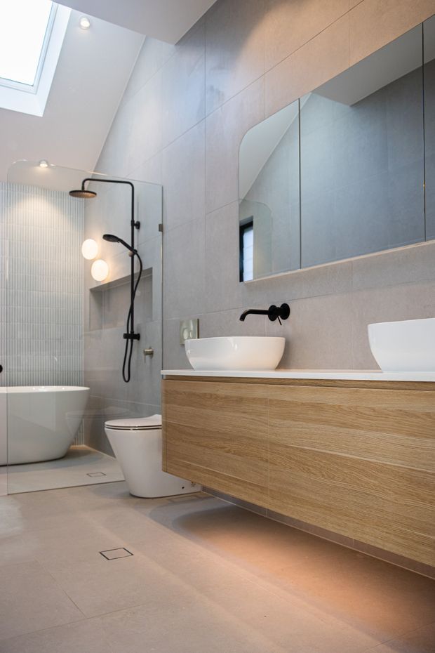 Contemporary bathroom with large-format light grey wall and floor tiles, a freestanding white bathtub, wall-mounted toilet, floating timber vanity with white vessel basins, black wall-mounted taps, frameless shower screen, and a large mirrored cabinet. A skylight and integrated lighting create a bright, refined atmosphere.