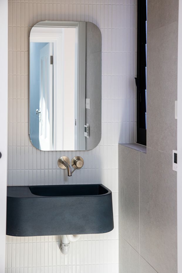A compact contemporary bathroom featuring a wall-mounted charcoal-grey basin with rounded corners, brushed brass wall-mounted tapware, a rounded-rectangle mirror, and vertically stacked narrow white wall tiles. A dark-framed window and grey stone-look wall surface add contrast.