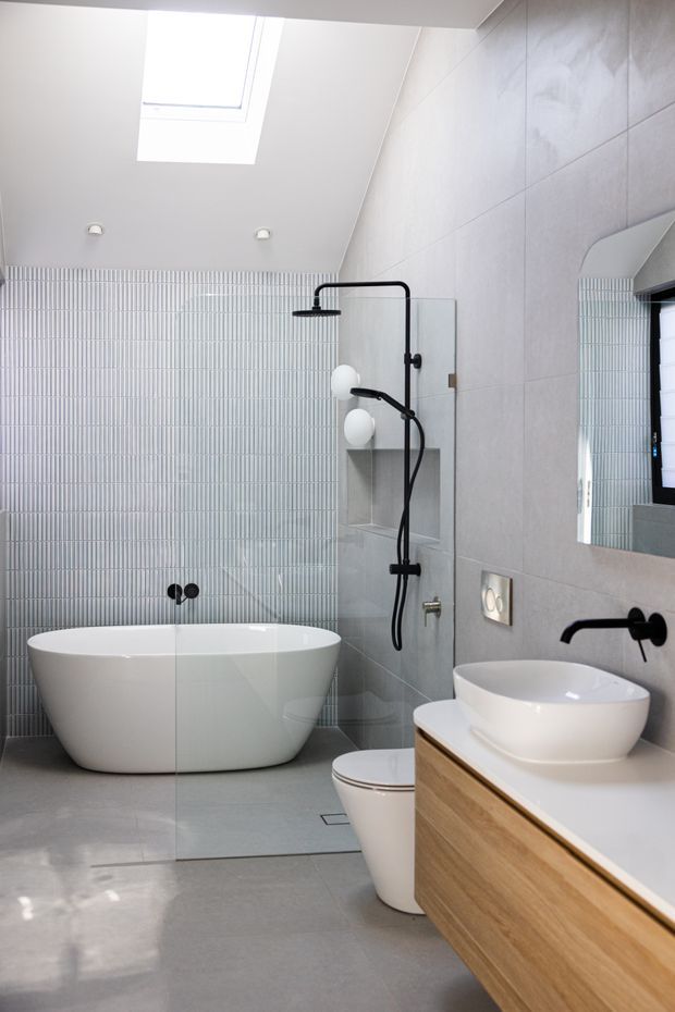Bright contemporary bathroom with a freestanding white bathtub, walk-in shower, vessel basin on a timber vanity, wall-hung toilet, large grey wall and floor tiles, and a vertically ribbed feature tile wall. Matte black tapware and shower fittings provide contrast, while a skylight brings in natural light.