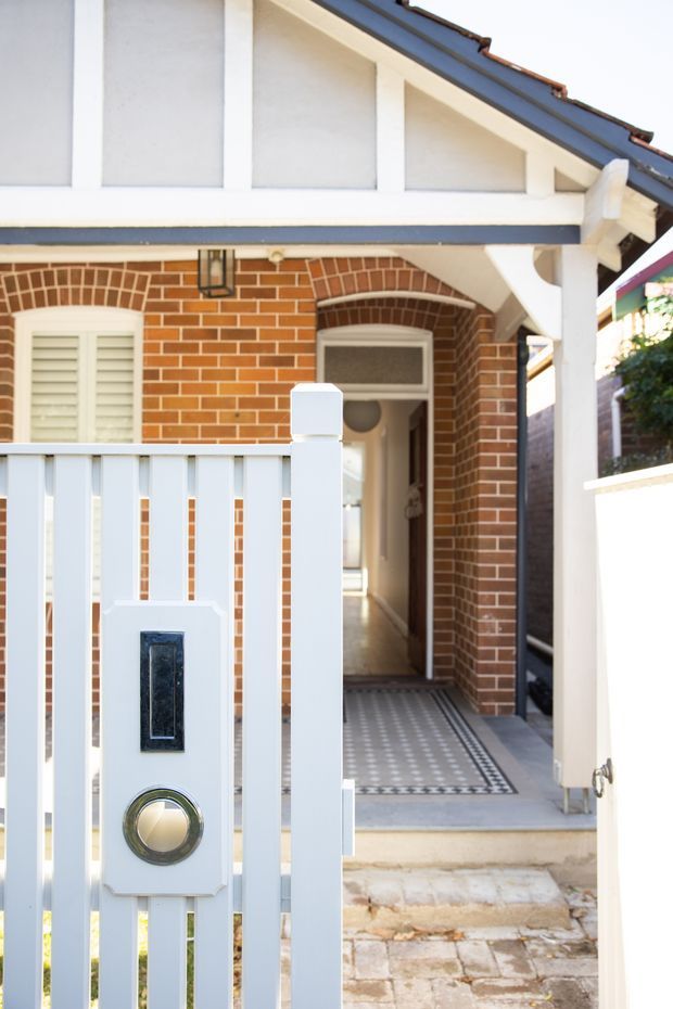 Street-level view of a traditional brick house entry with a white-painted timber front gate and fence, decorative intercom panel, tiled porch, arched doorway, louvred window shutters, and a covered gabled verandah with exposed decorative detailing.