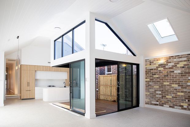 Bright contemporary open-plan interior with a white vaulted ceiling, exposed brick feature wall, pale timber kitchen cabinetry, white island bench, and large black-framed glazed openings. A double-height glazed volume connects to a small timber-decked outdoor courtyard, with a skylight bringing in additional natural light.