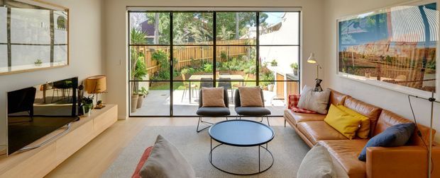 Bright contemporary living room with warm timber flooring, a low built-in media console, a tan leather sofa, lounge chairs, and a round black coffee table. Full-height black-framed glazed doors open to a leafy outdoor terrace or garden, while large framed artwork and a slim floor lamp add visual interest. The room has a relaxed, mid-century-influenced furnishing scheme with strong indoor-outdoor connection.