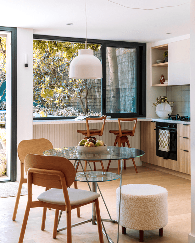 Bright contemporary kitchen-dining space with a compact timber kitchen, pale tiled splashback, large black-framed windows, and warm light timber flooring. A round glass dining table with timber chairs occupies the foreground, complemented by a sculptural pendant light and a small upholstered stool. The room has abundant natural light and views to planted outdoor greenery.
