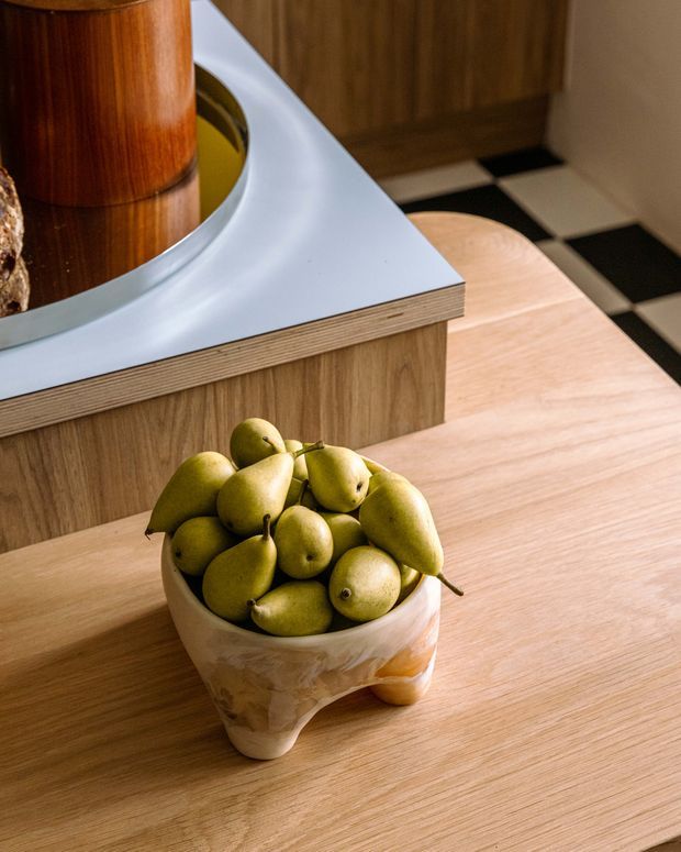 Close-up view of a warm-toned interior featuring a timber dining or kitchen work surface with a sculptural cream-colored bowl filled with pears. Behind it is a curved white benchtop with layered plywood edging, timber cabinetry, a cylindrical timber element, and a black-and-white checkerboard floor.