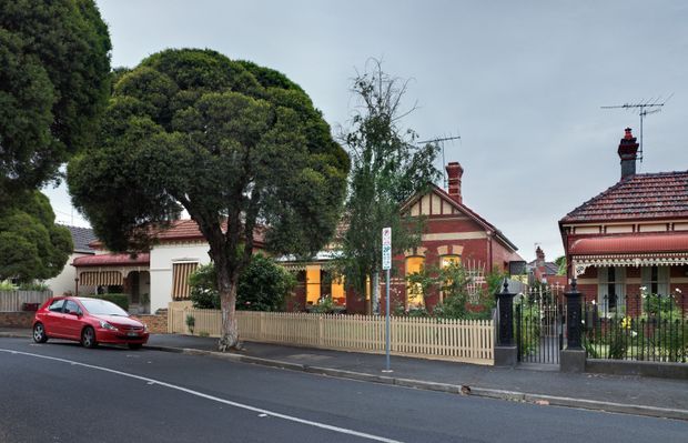 Street-level view of a traditional single-storey period residence in an established suburban streetscape. The house has a red tiled pitched roof, decorative gabled facade with timber detailing, red brick walls, cream-painted trim, verandahs with striped awnings, a low picket fence, and an ornate black metal front gate. Mature street trees partly screen the property.