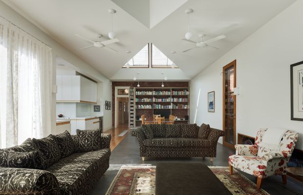 Bright open-plan living room with patterned sofas, a floral armchair, a large dark ottoman, and layered rugs. The room opens toward a compact white kitchen and a dining/library area with extensive built-in book shelving. A high pitched ceiling features triangular clerestory glazing, exposed timber framing, recessed lighting, and multiple white ceiling fans. Timber flooring, white walls, sheer curtains, and framed artwork create an airy, eclectic interior.