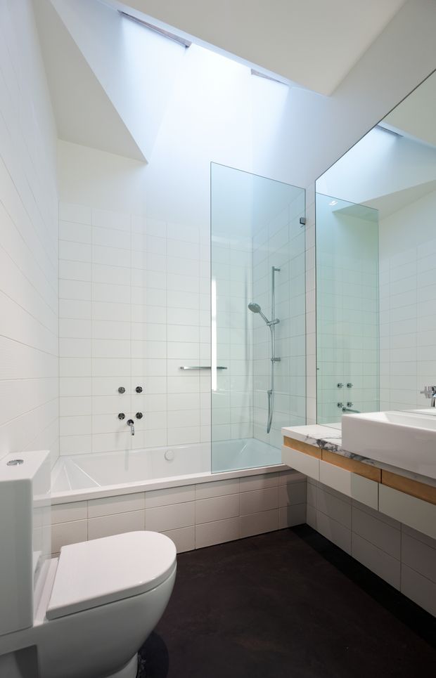 Bright contemporary bathroom with a white tiled bath and shower enclosure, wall-mounted shower fittings, a large frameless mirror, white vanity basin with timber cabinetry and a pale stone benchtop, plus a white toilet. A high angled ceiling and skylight-like glazing bring in abundant natural light, while the dark floor provides contrast.