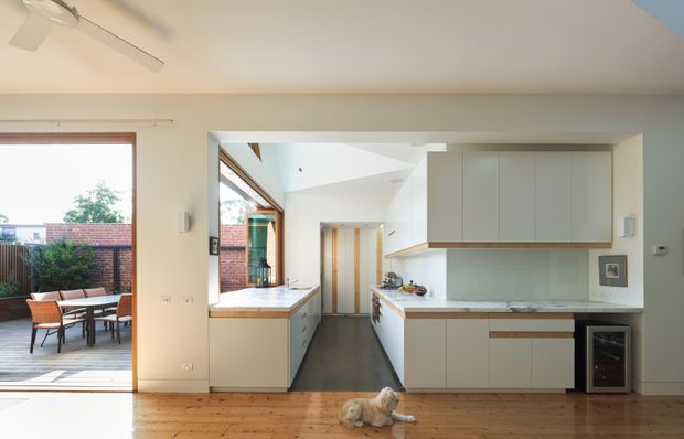 Bright open-plan kitchen viewed from an adjoining living area, with pale built-in cabinetry, a light stone benchtop, timber flooring, and a peninsula or island workspace. Large glazed doors open to a furnished outdoor terrace, while a skylit or vaulted opening brings additional daylight into the kitchen. A small dog is visible on the floor.