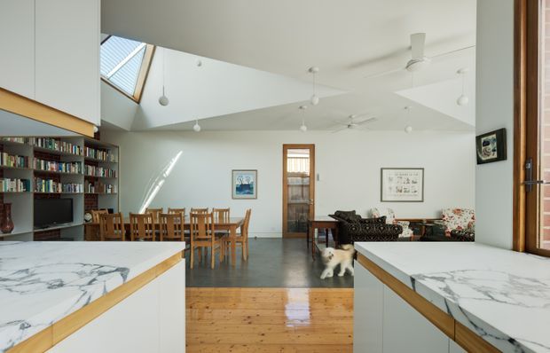 Open-plan residential interior viewed from a bright white kitchen, with marble-look island benchtops and timber edging in the foreground. The space combines a dining area and living room, featuring built-in dark timber bookshelves, a television, timber dining table and chairs, patterned sofa seating, polished timber flooring transitioning to a darker hard floor, a high angled ceiling with skylights, pendant lights and ceiling fans, and a glazed timber door. A small dog is present in the living area.