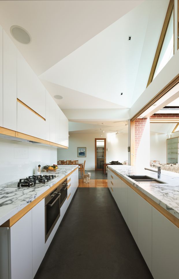 Bright contemporary galley kitchen with extensive white flat-panel cabinetry, dramatic white marble-patterned benchtops, integrated stainless-steel appliances, and a dark flooring aisle. A timber dining area and living space extend beyond the kitchen, while a large glazed opening and exposed brick wall bring natural light and texture into the room. The high, angular ceiling incorporates recessed lighting and a skylight-like clerestory opening.