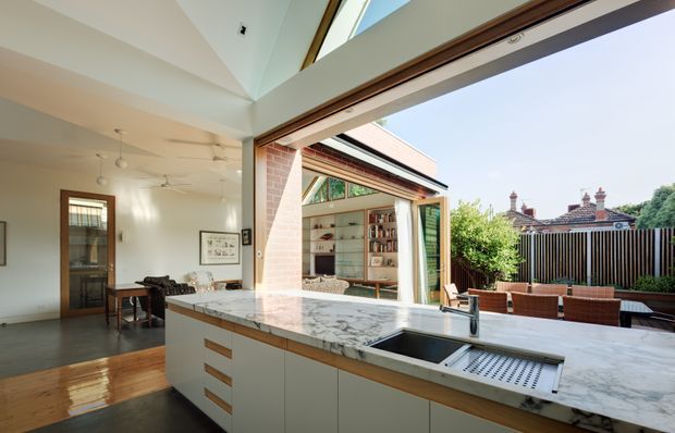 Bright open-plan kitchen viewed from inside, with a large marble-look island, integrated sink and drainer, pale cabinetry with timber drawer accents, and wide open glazing/doors leading to a brick-paved outdoor terrace. The adjoining living/dining area and built-in shelving are visible, along with a high angled ceiling and clerestory glazing. The scene has a warm contemporary renovation character with strong indoor-outdoor connection.