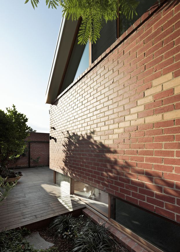 Exterior architectural view of a brick-clad residential building, showing a tall red brick wall with contrasting pale brickwork, deep roof overhang, vertical glazing at the lower level, and a timber deck walkway beside planted garden beds. Strong sunlight casts foliage shadows across the facade.