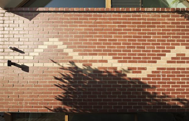 Close-up exterior view of a red brick wall with contrasting pale brick sections forming a subtle geometric pattern. A dark horizontal coping course runs along the top, with black wall-mounted exterior lights and strong tree shadows across the facade. A small portion of dark glazing or an opening is visible at the base.