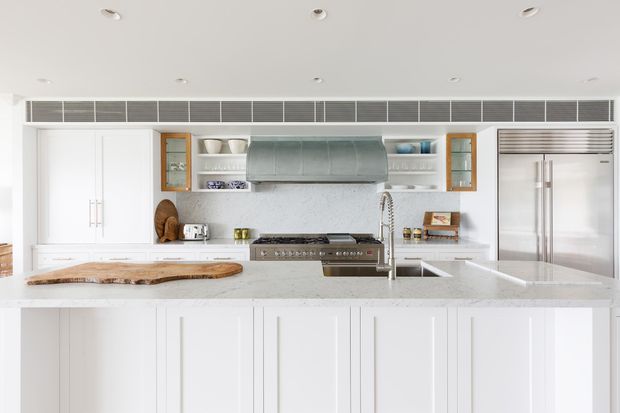 Bright, spacious contemporary kitchen with extensive white cabinetry, a large central island, integrated stainless-steel appliances, open shelving, glass-front timber-accented cupboards, and a prominent curved metal rangehood. The island includes a pale stone benchtop and a contrasting live-edge timber board. Recessed ceiling lights and a long linear ventilation grille contribute to the clean, refined fit-out.