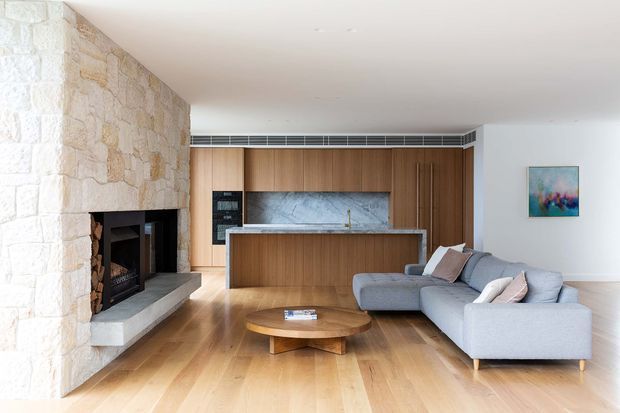 Bright contemporary open-plan living room with a large light-stone fireplace wall, built-in timber kitchen cabinetry and island, pale stone splashback, timber flooring, and a light grey sectional sofa. The room has a clean, refined fit-out with warm natural materials and recessed lighting.