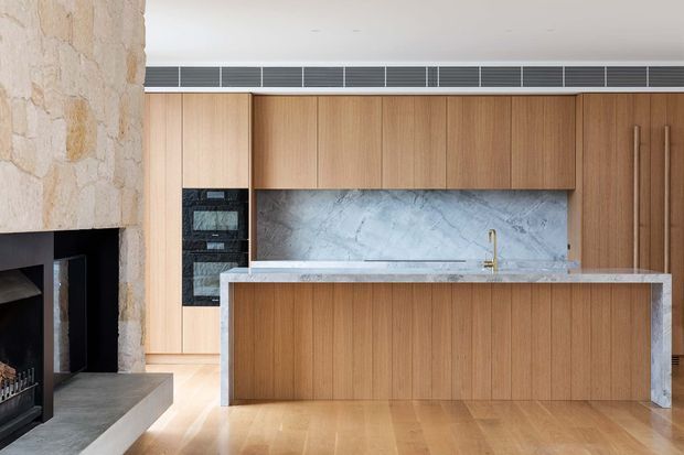 Contemporary open-plan kitchen with full-height light timber cabinetry, a large central island with a pale stone benchtop, and a matching marble-look stone splashback. A textured natural stone fireplace wall and dark built-in fireplace are visible at the left. The space has warm timber flooring, integrated appliances, slim linear ventilation, and restrained modern detailing.