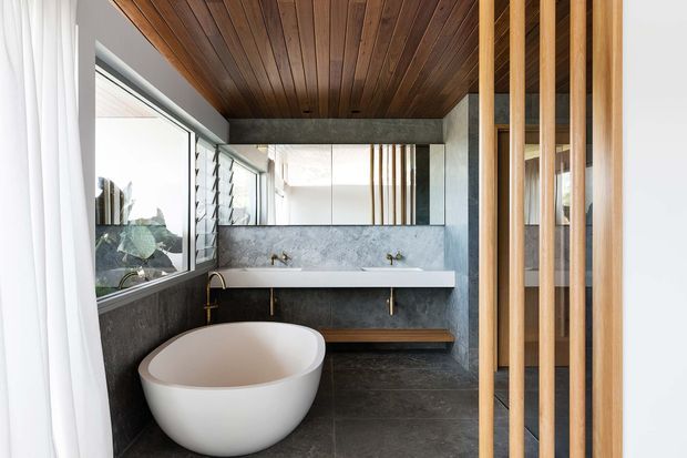 A contemporary bathroom with a freestanding white oval bathtub, a long double-basin wall-mounted vanity, dark stone-look wall and floor finishes, brass fixtures, timber ceiling panelling, and a vertical timber slat screen. Large windows and mirrors bring in abundant natural light.