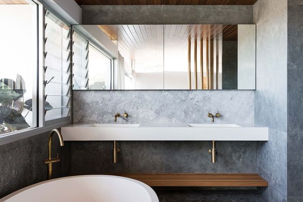 Contemporary double-vanity bathroom with a long white stone or solid-surface counter, wall-mounted basins and taps, a large frameless mirror, dark grey stone-look wall finishes, warm timber ceiling and bench, and extensive louvred glazing along the left side. A freestanding white bathtub is partially visible in the foreground.